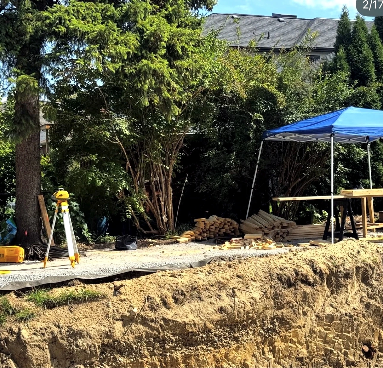 Real Pisces jobsite: surveyor laser level on tripod beside lumber stacks at the edge of an excavated foundation pit